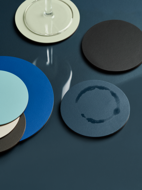 View of a blue glass table featuring seven linoleum PAD coasters in cool-toned colours, with a glass ring mark on one coaster and the reflection of a wine glass visible on the table